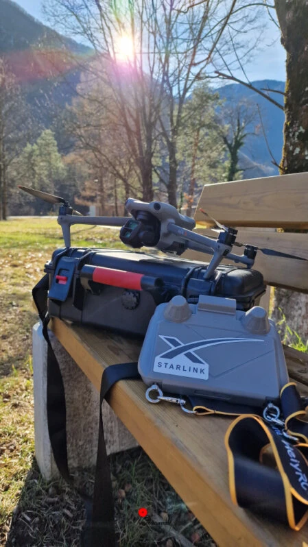 Attrezzatura Starlink e drone su panchina in montagna Kit Starlink e drone posizionati su una panchina in legno con sfondo montano e luce solare filtrata tra gli alberi